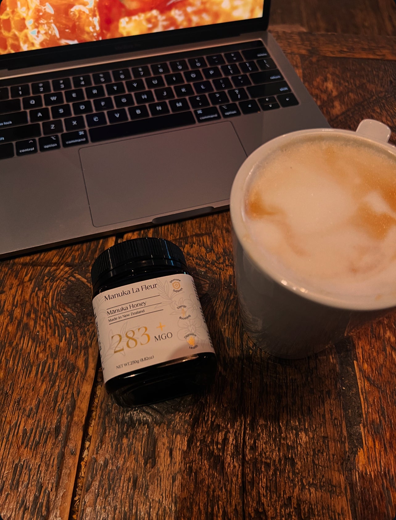 Laptop, bottle of Manuka Honey, and cup of coffee on a wooden table.